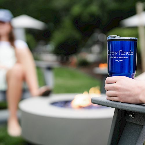 A person holds a blue Greyfinch cup near a fire pit while another person sits in the background. Outdoor setting with blurred greenery around.