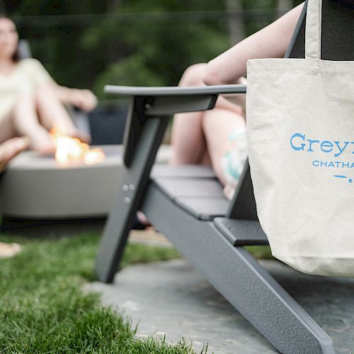 A group of people sitting around a fire pit outdoors with a tote bag hanging off a chair that reads "Greyfinch Chatham Inn."