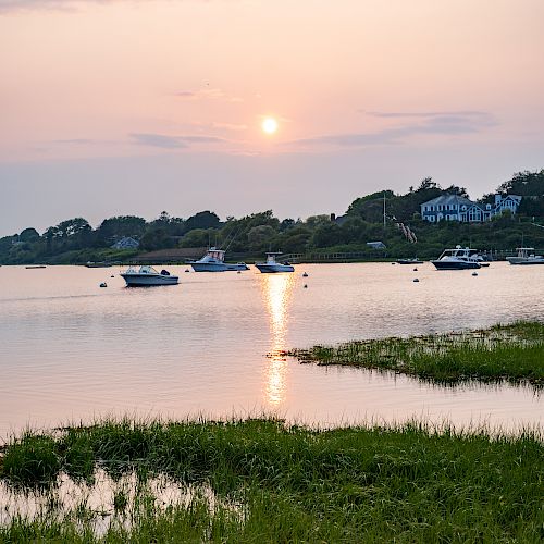 A serene sunset over a calm lake, with a few boats anchored near the shore and lush greenery in the foreground.