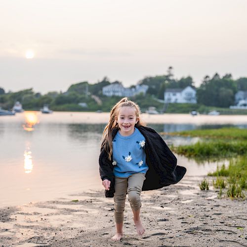 A young girl is barefoot on a beach, running and smiling at the camera, with boats on the water and houses in the background.