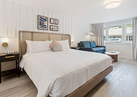 A modern, well-lit bedroom with a large bed, nightstand, wall art, sofa, small table, and a window with curtains, featuring light-colored decor.