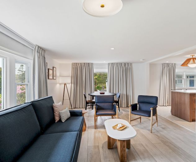 A modern living room with a navy blue sofa, wooden coffee table, armchairs, and a dining area, with large windows and light wooden flooring.