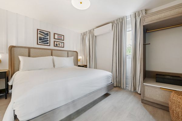 A minimalistic bedroom with a large white bed, wall art, a wardrobe, and curtains covering a window.