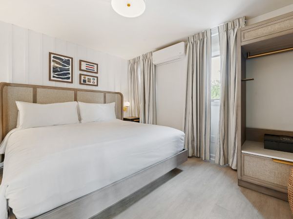 A minimalistic bedroom with a large white bed, wall art, a wardrobe, and curtains covering a window.