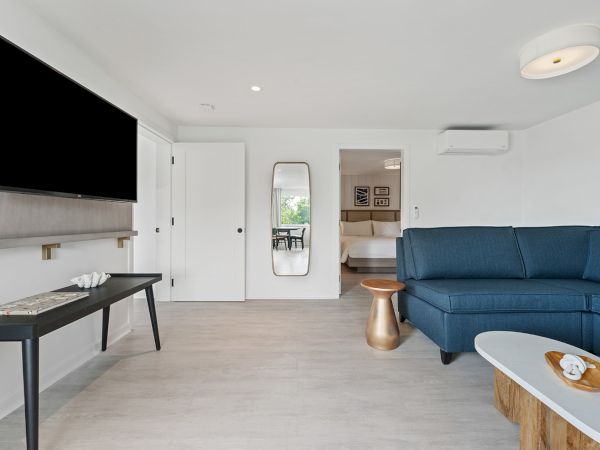 A modern living room with a large TV, blue sectional sofa, wooden table, black console table, and a full-length mirror. A bedroom is visible in the back.