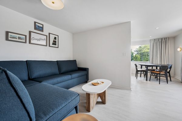 A modern living area with a blue sofa, wooden coffee table, picture frames on the wall, leading to a dining area with a table and chairs by the window.