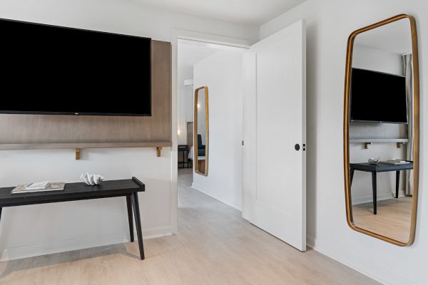 A modern room with a wall-mounted TV, a black desk, an open door, and a mirror reflecting another TV and part of the room next door.