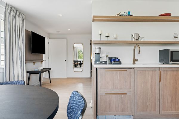 A modern room with a kitchenette, dining table, and TV. The kitchenette has a microwave, coffee maker, shelves, and a sink with a gold faucet.