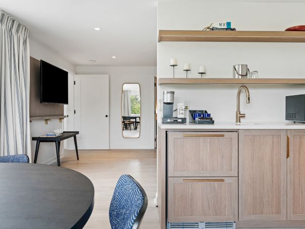 A modern room with a kitchenette, dining table, and TV. The kitchenette has a microwave, coffee maker, shelves, and a sink with a gold faucet.