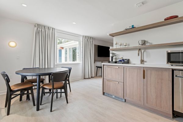 This image shows a modern kitchen and dining area with a round table and four chairs, open shelving, a microwave, and a wall-mounted TV.