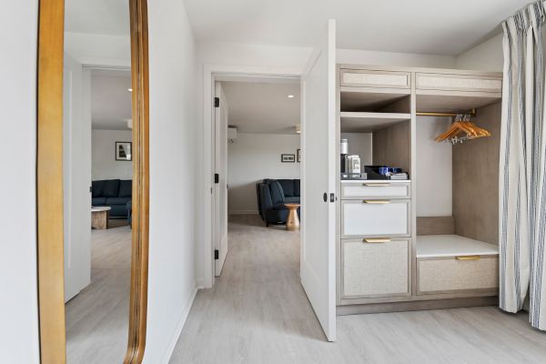 A modern room features an open wardrobe, a mirror on the left, and a view into a living area with blue furniture through an open door.