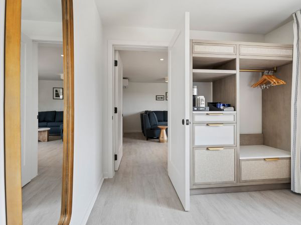 A modern room features an open wardrobe, a mirror on the left, and a view into a living area with blue furniture through an open door.