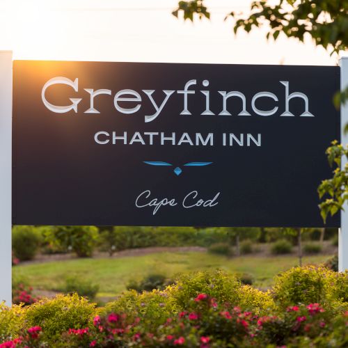 The image shows a sign reading "Greyfinch Chatham Inn Cape Cod," surrounded by greenery and flowers, suggesting it is near a landscaped area.