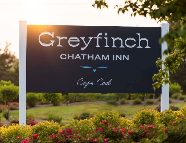 A large outdoor sign reads &ldquo;Greyfinch Chatham Inn&rdquo; with &ldquo;Cape Cod&rdquo; script below, set among greenery and flowers at sunset.
