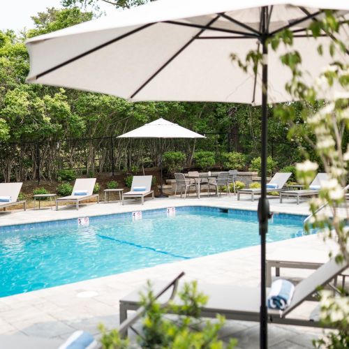 A sunny poolside scene with lounge chairs, umbrellas, and a clear blue swimming pool surrounded by greenery.
