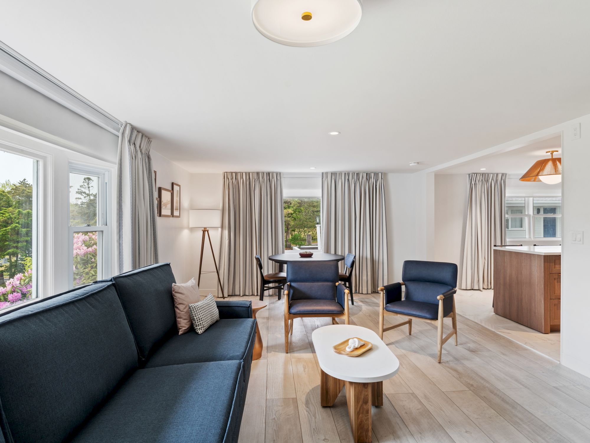 A modern living room with a navy blue sofa, matching chairs, a white coffee table, and large windows with beige curtains, adjacent to a dining area.