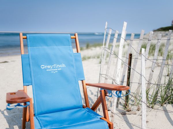 A blue beach chair with "Greyfinch Chatham, MA" print is on a sandy beach near a fence, with ocean waves in the background.