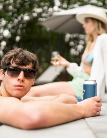 A person relaxes by the poolside in sunglasses, holding a can, with another person in the background wearing a hat and swimsuit.