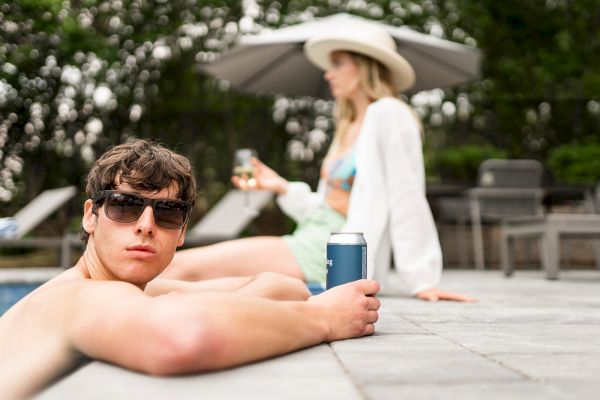 A person in sunglasses relaxes by a pool holding a drink, with another person in a hat and swimsuit seated in the background.