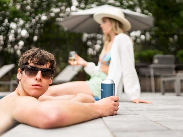 A person in sunglasses relaxes by a pool holding a drink, with another person in a hat and swimsuit seated in the background.