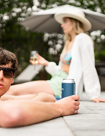 A man in sunglasses holds a can by a pool, with a woman in a hat and swimsuit sitting in the background.