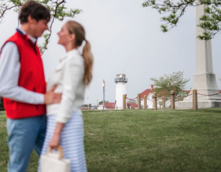 A couple stands together in a park with a lighthouse and some buildings in the background.