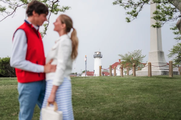 A couple stands together in a park with a lighthouse and some buildings in the background.