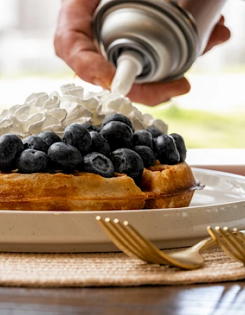 A waffle with blueberries and strawberries is being topped with whipped cream from a can, placed on a white plate with a woven placemat.