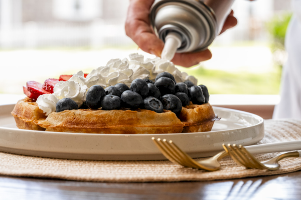 A waffle topped with blueberries and strawberries is being covered with whipped cream from a can, served on a plate with forks.