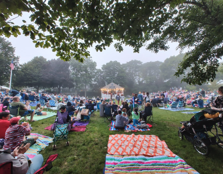 People are gathered on a lawn with blankets and chairs, watching a performance at a gazebo. Trees frame the scene.