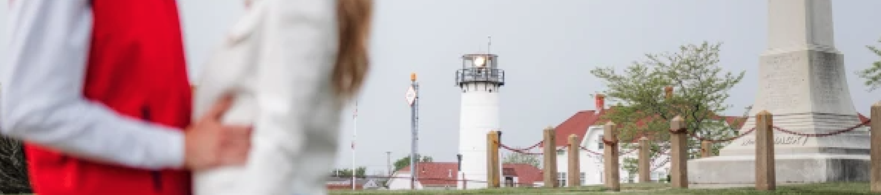 A couple stands closely, facing each other, with a lighthouse and monument in the blurred background on a grassy area.