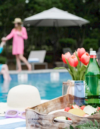 A poolside picnic scene with drinks and snacks on a tray by the pool while people lounge and chat in the background, sunny and relaxed.