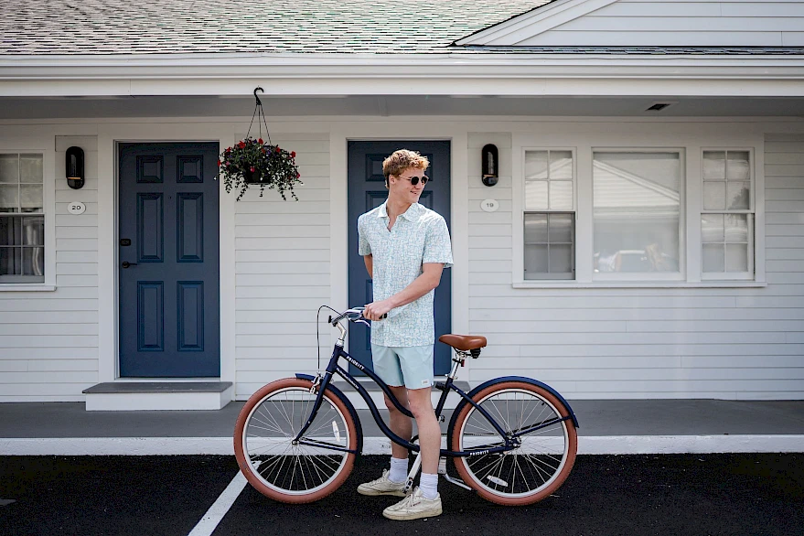 A person wearing light clothing stands next to a vintage bicycle with pale pink tires in front of a gray house with blue doors and hanging planter, on a sunny day.