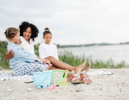 A mother with two kids sits on a beach, with a sandcastle bucket, beach tools, and sandals nearby by the water.
