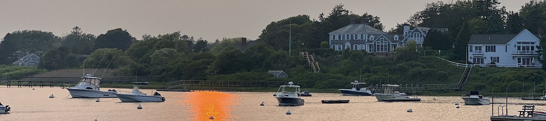 Sunset over a calm harbor with several boats anchored near a grassy shore and white houses reflecting in the water.