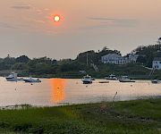Sunset over a calm harbor with several boats anchored near a grassy shore and white houses reflecting in the water.