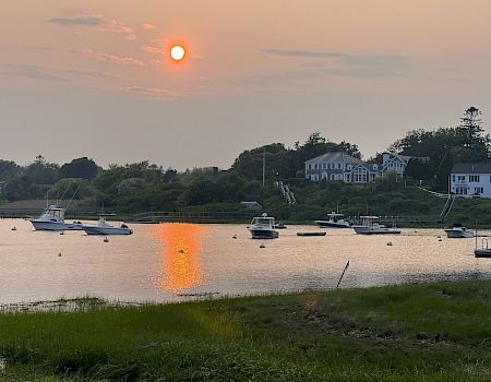 Sunset over a calm harbor with several boats anchored near a grassy shore and white houses reflecting in the water.