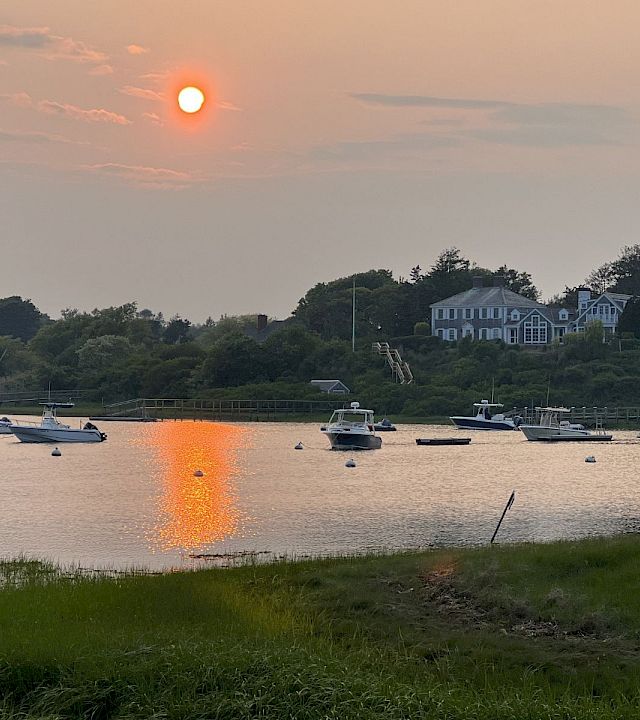 Sunset over a calm harbor with several boats anchored near a grassy shore and white houses reflecting in the water.