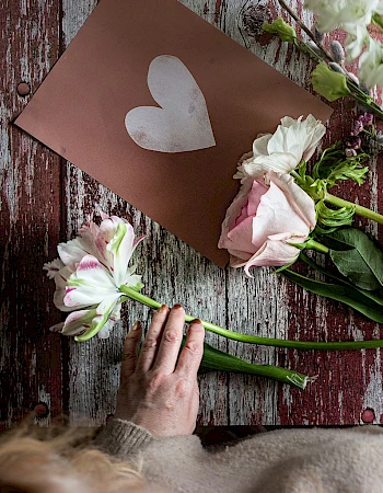 A person arranges pink and white roses on a rustic wooden surface, placing stems next to a card with a white heart. End sentence.