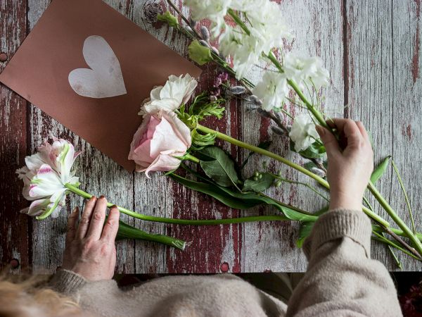 A person arranges pale pink roses and white flowers on a rustic wooden surface, with a brown card featuring a white heart.