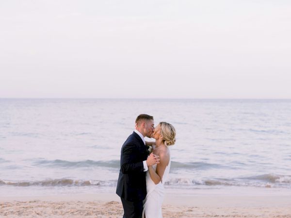 A couple in wedding attire embrace and kiss on the beach, gentle waves in the background, serene sunset vibes, romantic moment.