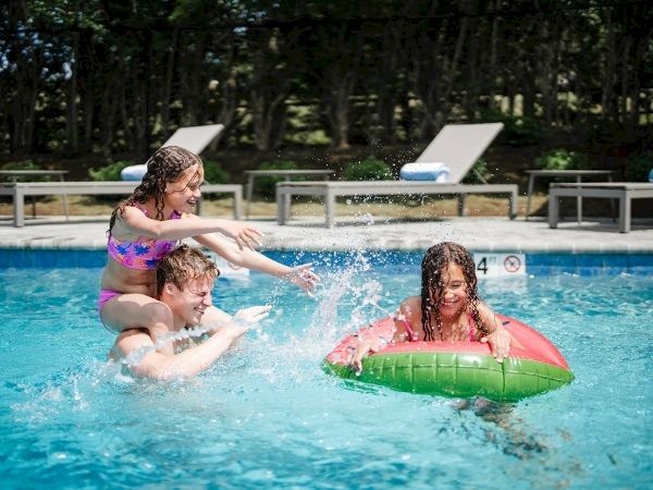Three kids play in a sunny pool; one girl in a pink swimsuit helps another near the edge while a third girl lounges on a green inner tube.