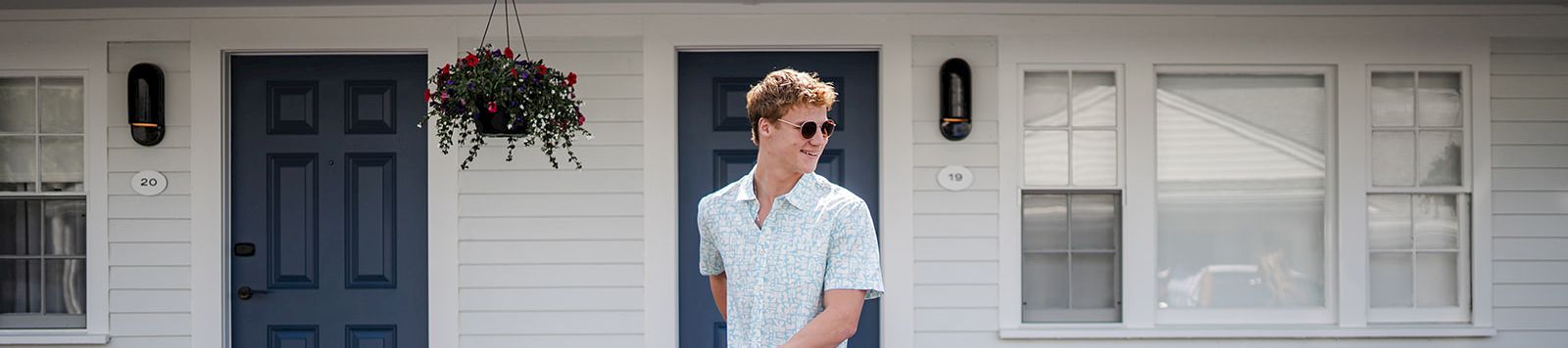 A man in light blue outfits stands with a vintage bicycle in front of a white house with blue doors; hanging plant, sunny day.