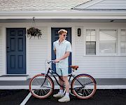 A man in light blue outfits stands with a vintage bicycle in front of a white house with blue doors; hanging plant, sunny day.