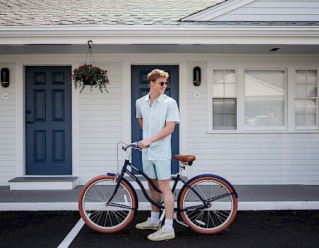 A man in light blue outfits stands with a vintage bicycle in front of a white house with blue doors; hanging plant, sunny day.