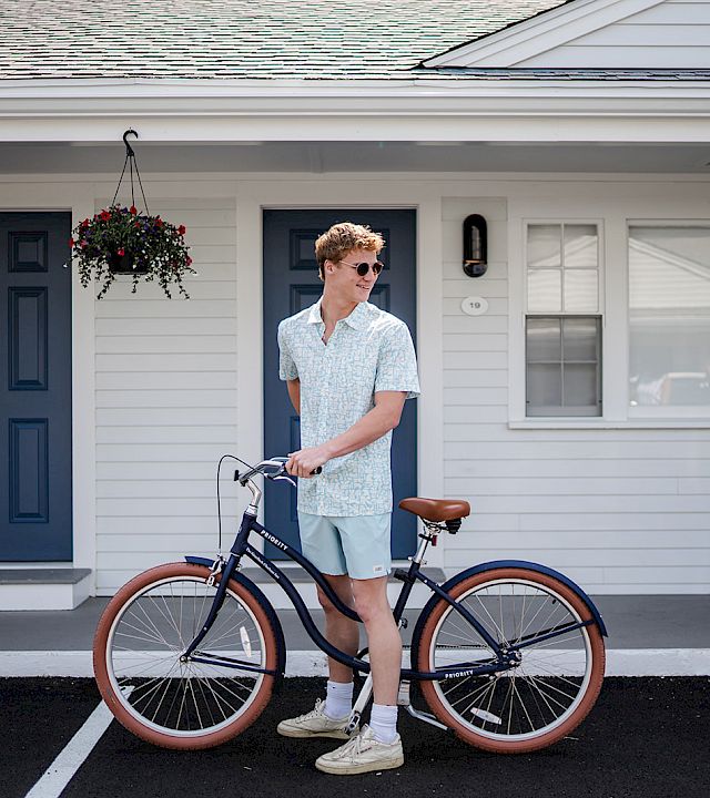 A man in light blue outfits stands with a vintage bicycle in front of a white house with blue doors; hanging plant, sunny day.