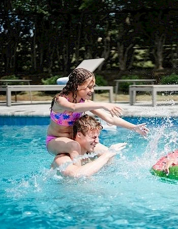 Kids having fun at a sunny pool: two girls push a boy underwater while another girl floats on a green inner tube, all splashing and laughing.