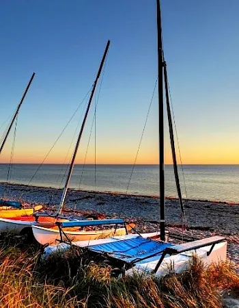 A sailboat rests on a grassy beach at sunset, with waves and warm sunlight on the ocean.