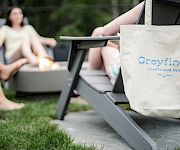 A beige tote with “Greyfinch” logo rests on a lounge chair in a sunny backyard, while people sit around a fire pit chatting.