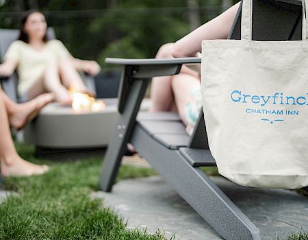 A beige tote with &ldquo;Greyfinch&rdquo; logo rests on a lounge chair in a sunny backyard, while people sit around a fire pit chatting.